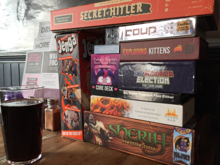 A stack of board games on a table, including Secret Hitler, Coup, Exploding Kittens, Twilight Sparkle's Secret Shipfic Folder, Elections of US America Election: The Card Game, We Didn't Playtest This Either, Flame War, Sheriff of Nottingham, and Jenga. A partially drank pint of cola stands next to the games.