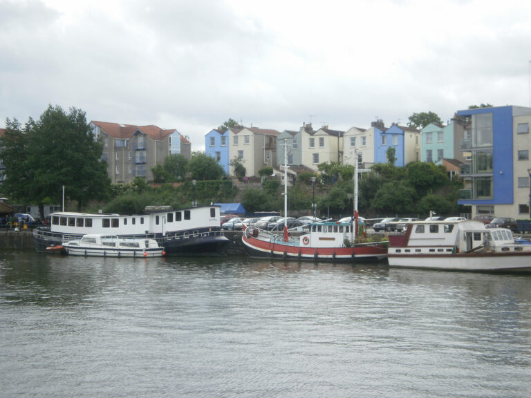 Several large boats parked along the side of a harbour. Tall, narrow, brightly painted houses rise up from behind them.
