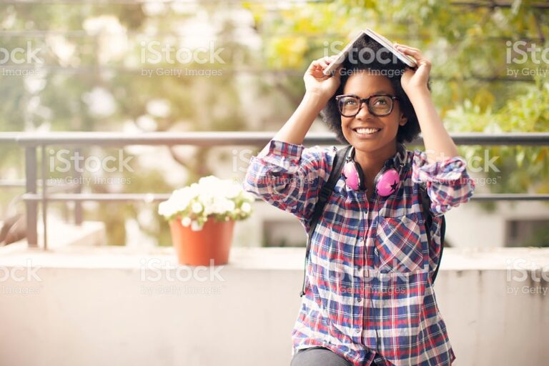 A young black woman wearing glasses, backpack and pink headphones sits outside, smiling and holding a book over her head. The image is covered in iStock/Getty Images watermarks, for the aesthetic.