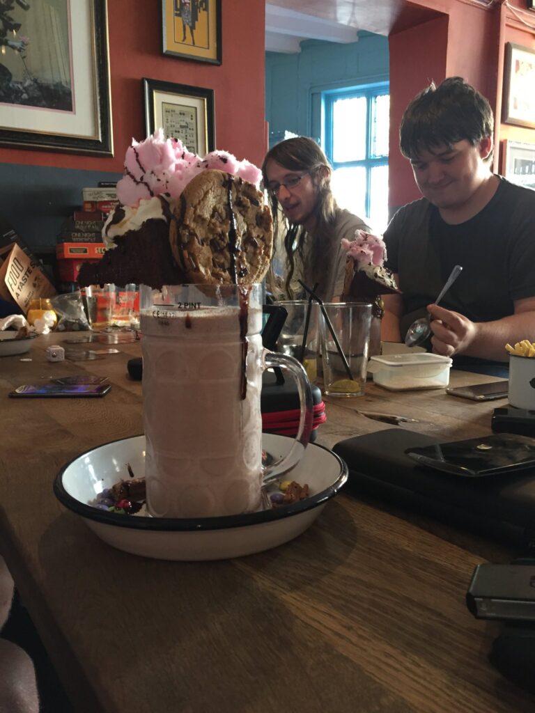 A photo of an insane dessert, composed of a large glass containing two pints of chocolate milkshake, on top of which are balanced a large chocolate chip cookie, a slice of chocolate cake, and a bunch of pink candy floss. The glass is surrounded by Nutella spread and M&Ms, and the entire thing is covered in chocolate sauce.
