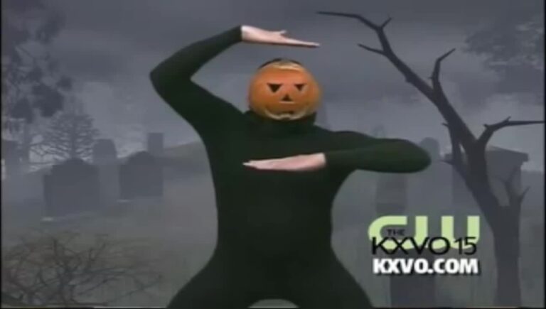 A person dressed in a black bodysuit, with a jack-o'-lantern covering their face, dancing in front of a green screened image of a graveyard.