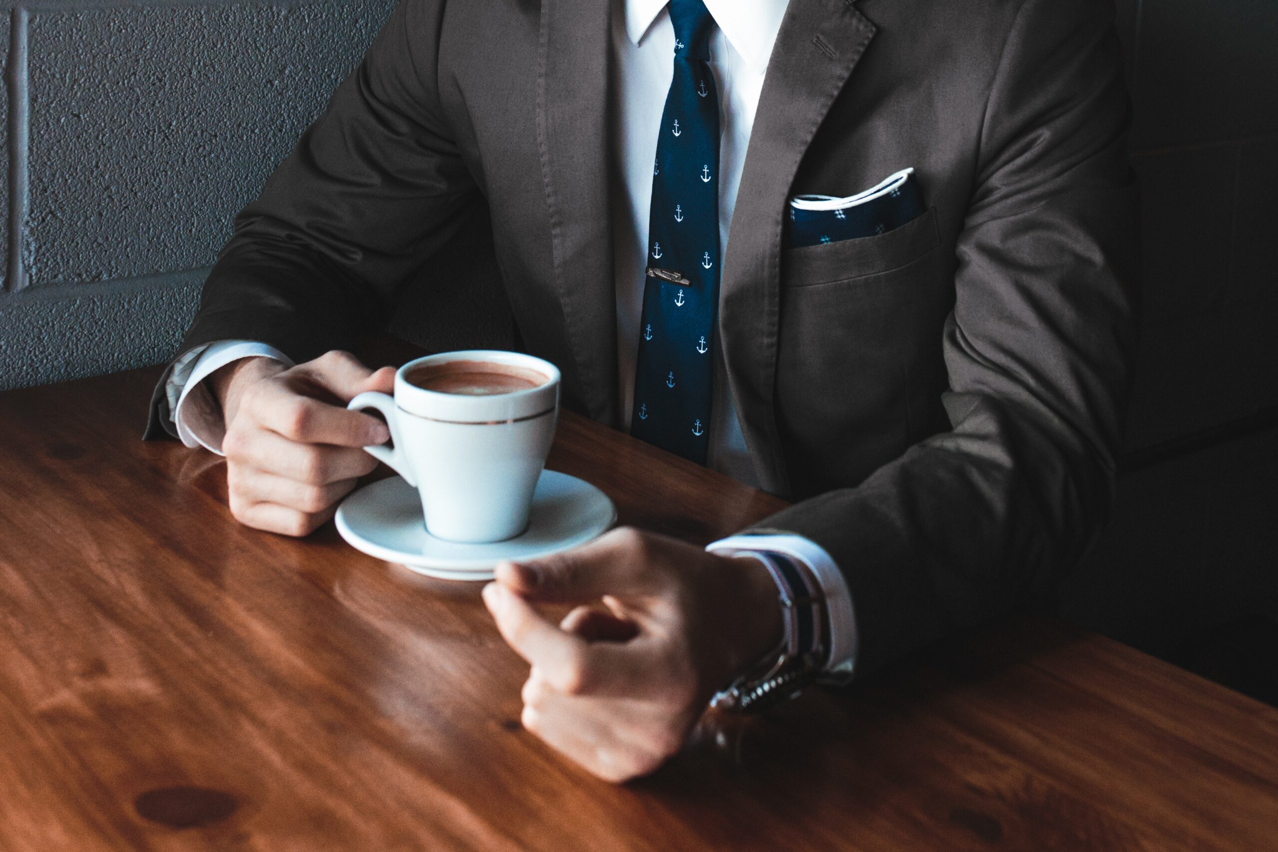 A close up of a man wearing a business suit sat at a table drinking coffee.