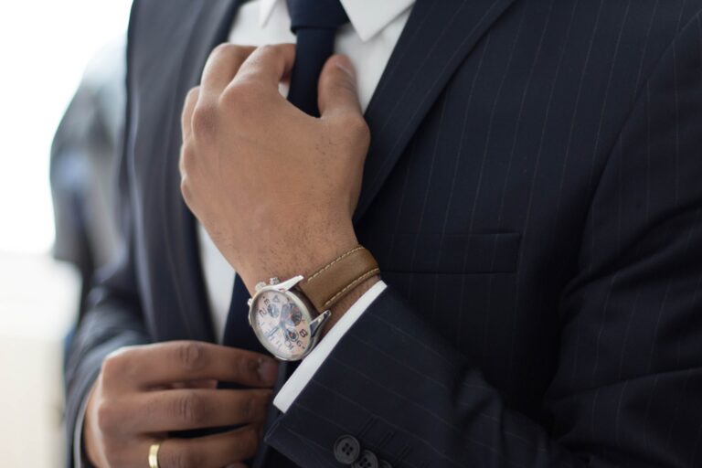 A close up of a man wearing a business suit's upper torso. A gold ring and wristwatch is clearly visible as he adjusts his tie.